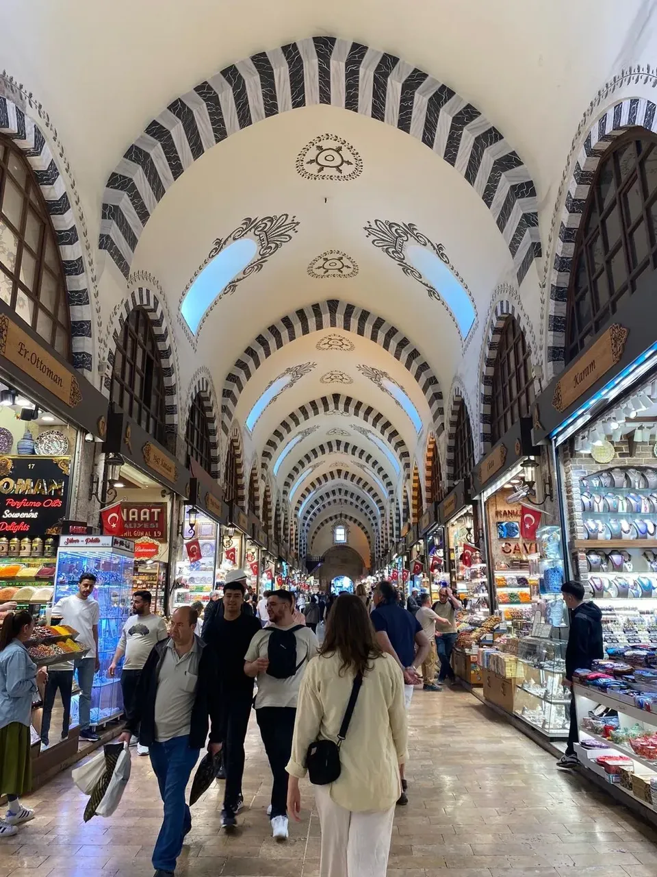 The bustling Grand Bazaar in Istanbul, filled with a variety of stalls selling goods like textiles, lanterns, jewelry, and ceramics, with crowds of people walking through.