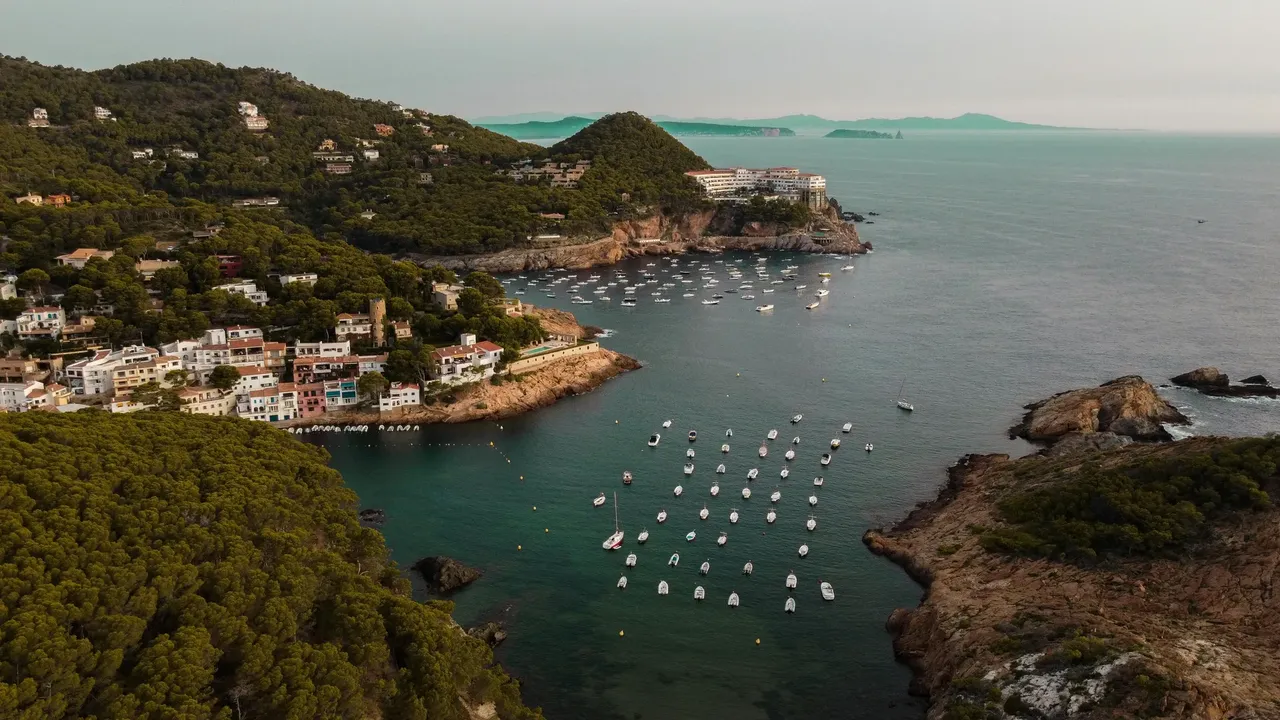 O priveliște pitorească a litoralului Costa Brava, cu țărmuri stâncoase, ape albastre clare și vegetație luxuriantă.