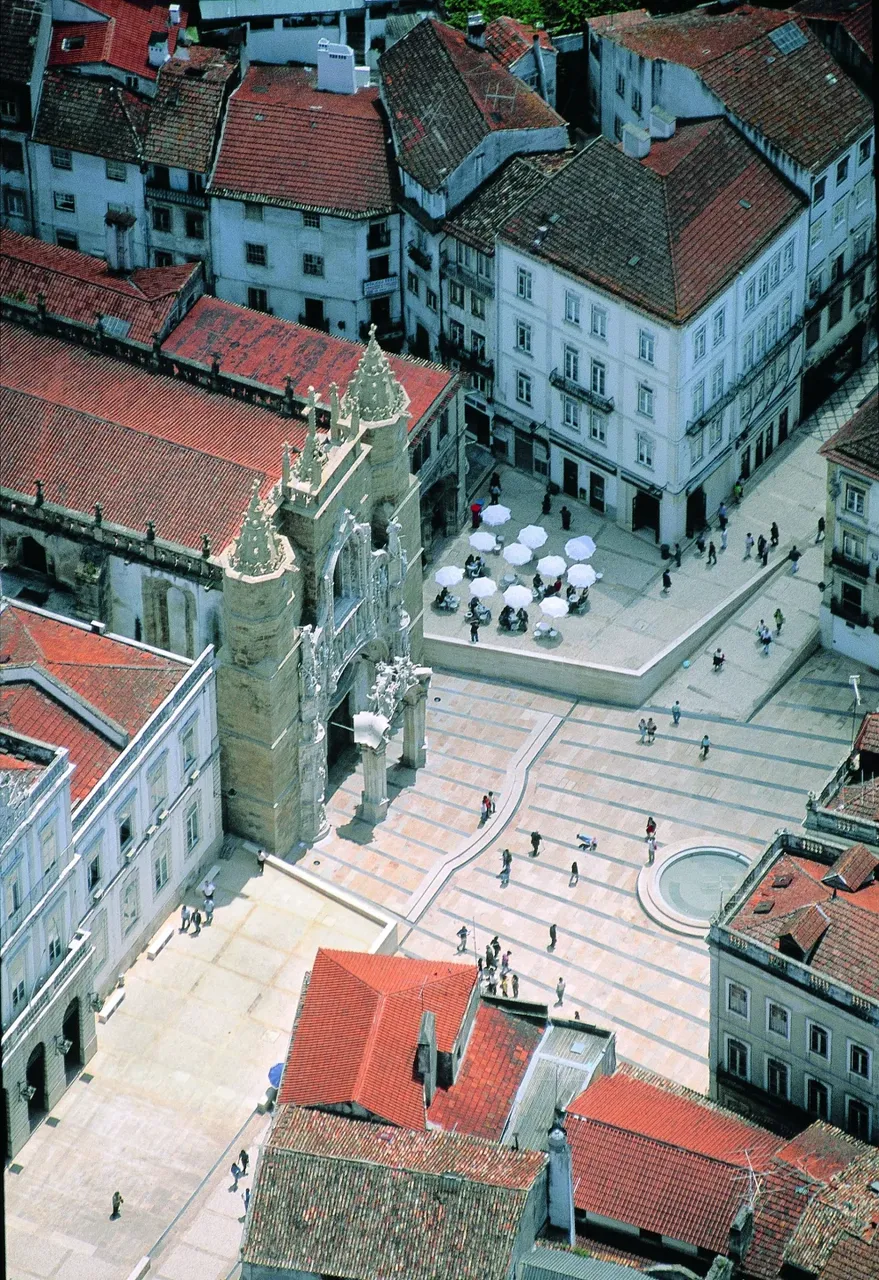 Aerial view of Praça 8 de Maio in Coimbra, Portugal, featuring the ornate Santa Cruz Church, patterned stone plaza, red-tiled rooftops, and people walking and sitting at outdoor café tables.