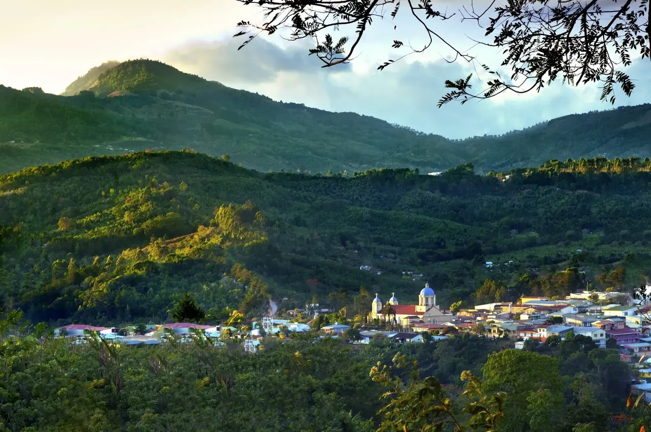 A panoramic aerial view of San José, Costa Rica, nestled in a lush valley between towering green mountains, with the city sprawling out below.