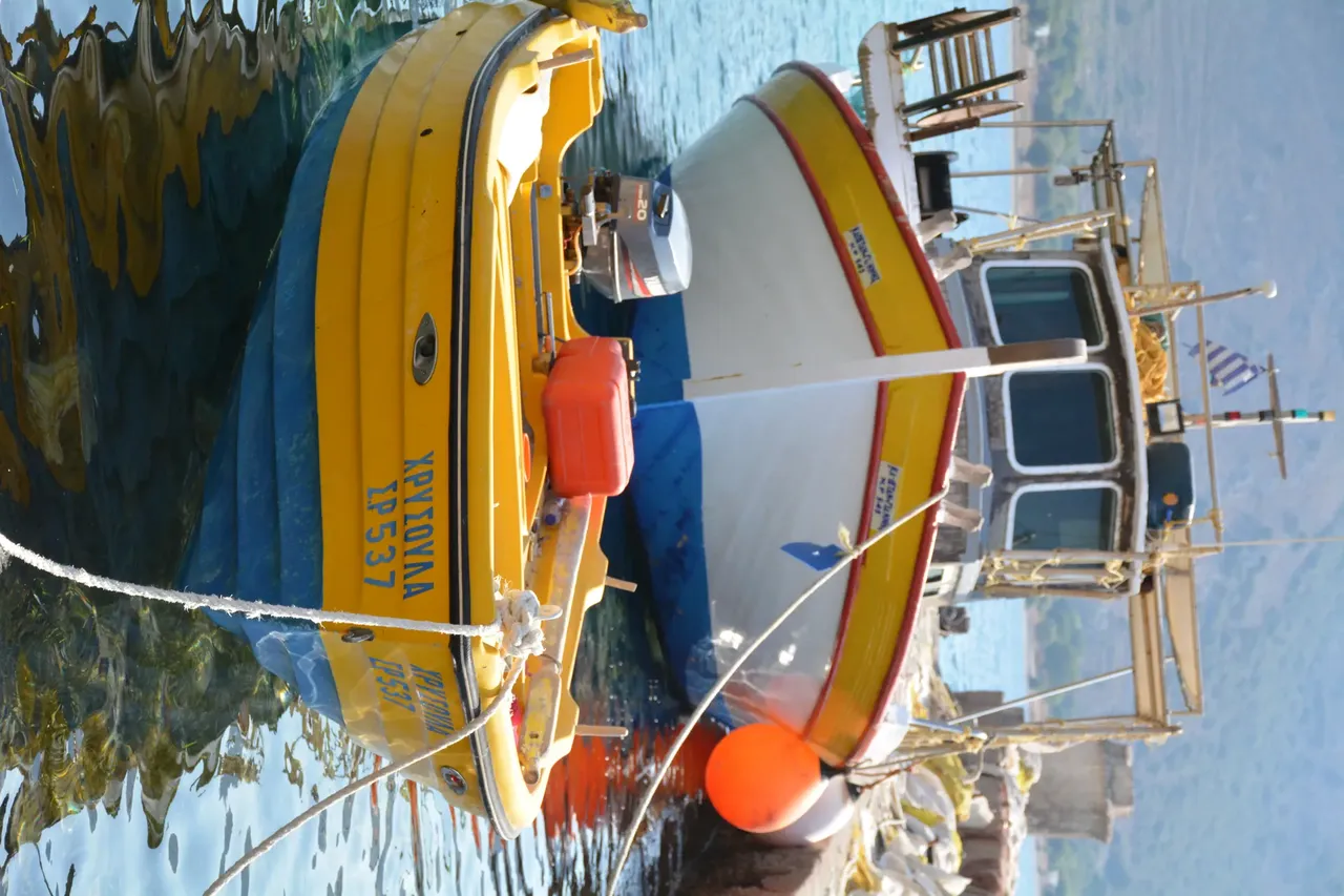 Two small boats moored at a serene harbor in Tilos, Greece, with clear blue waters reflecting the surrounding landscape and a hilly backdrop
