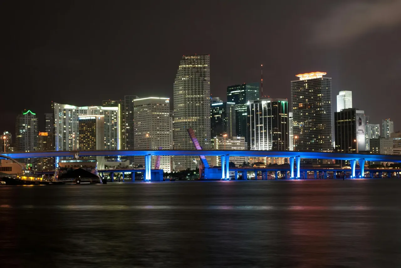 The vibrant skyline of Miami, Florida, featuring tall, modern skyscrapers and colorful lights reflecting off the water at dusk.