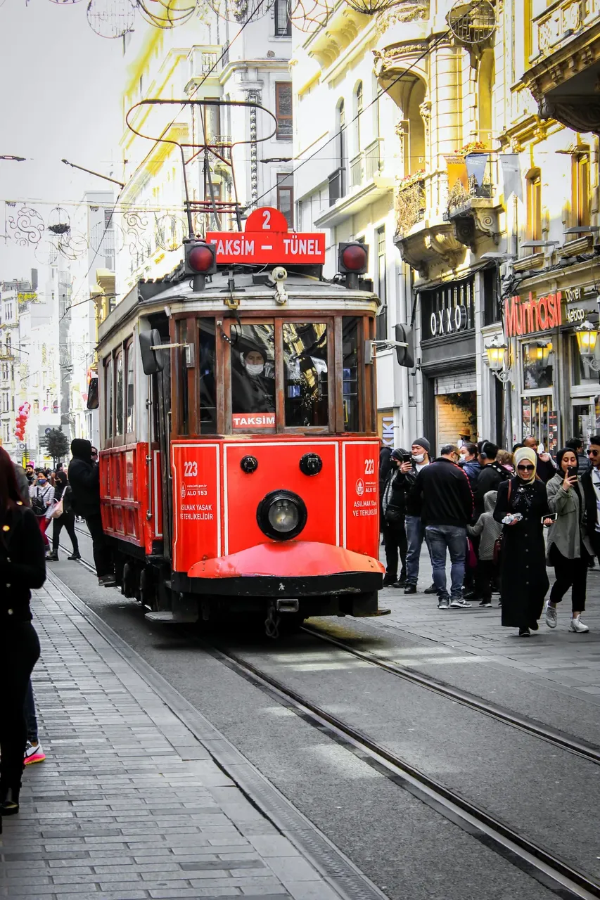 The historic red tram in Taksim Square, Istanbul, moving along a busy street with pedestrians and shops lining the sidewalks.
