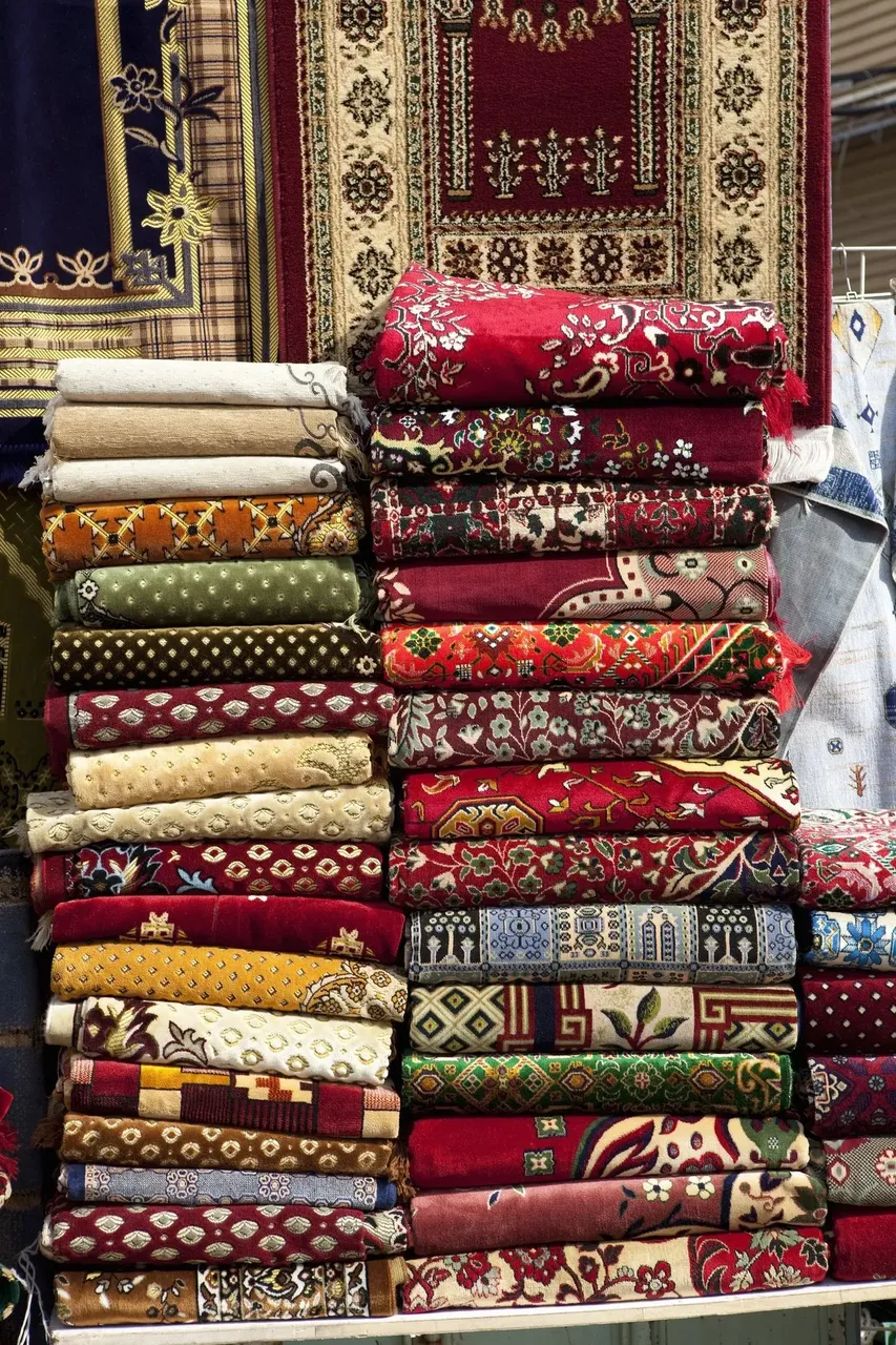 ibrant, intricately designed carpets displayed in stacks and rows at the Souk Al Alawi, showcasing traditional Middle Eastern patterns.