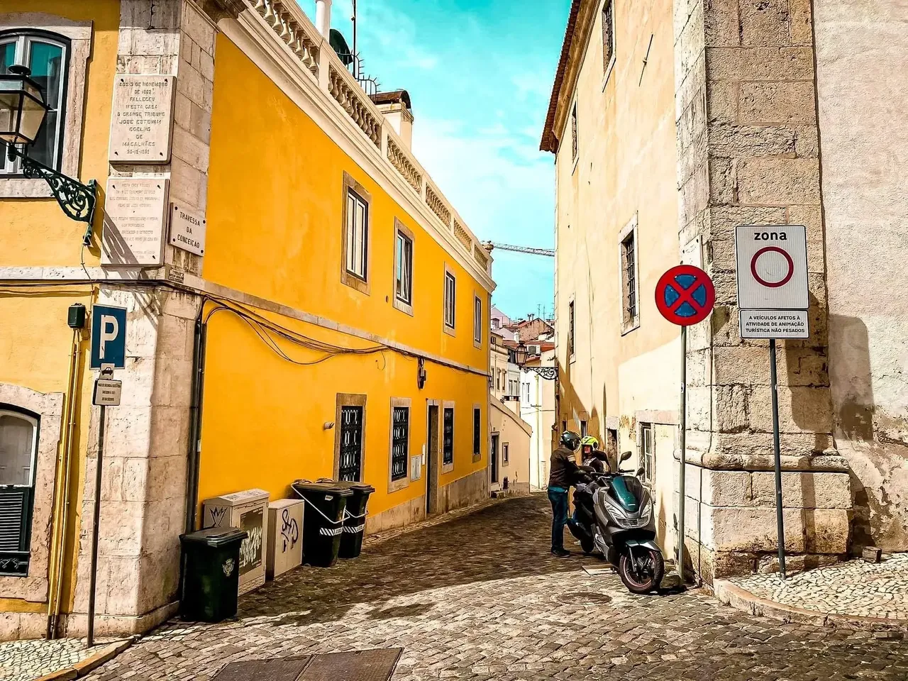 Bright yellow and beige buildings lining a cobblestone street in Lisbon, Portugal, with a parked scooter and street signs