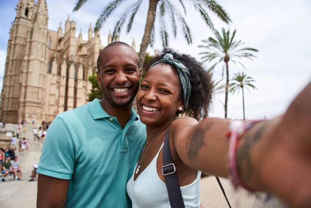 young couple taking a selfie on their holidays in Spain, looking at camera with big toothy smile. Huge cathedral church in the background.