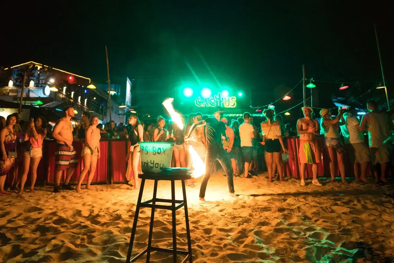 Bulizók táncolnak és élvezik a lüktető éjszakai Teliholdpartit Ko Phangan strandjának színes fényei alatt.