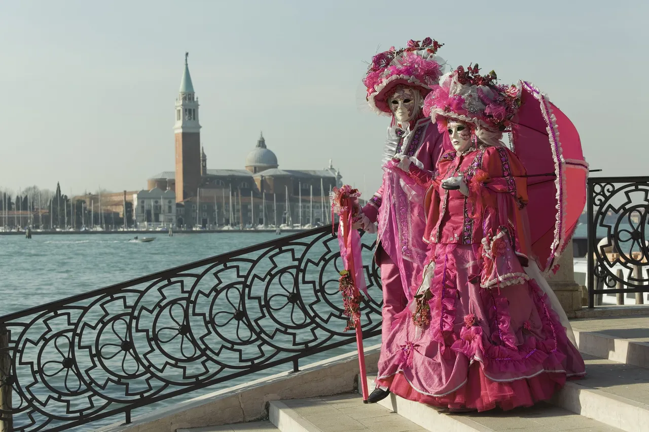 Two elaborately dressed individuals in traditional Venetian carnival costumes and masks, posing with the iconic view of a plaza across the river in Venice.