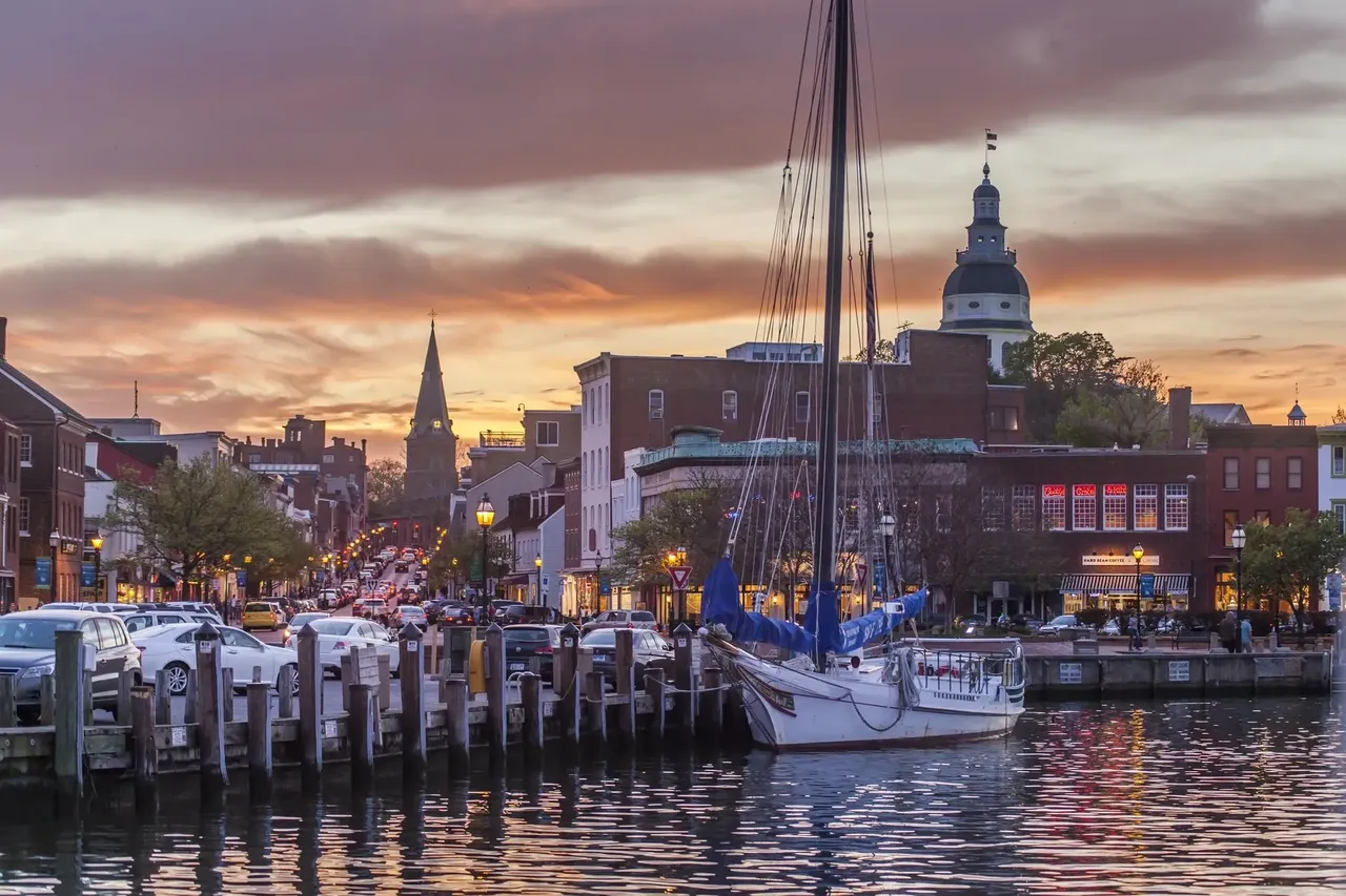 A Maryland állambeli Annapolis kikötőjének nyugodt látványa, a vízen finoman úszó vitorlásokkal, a vízparton álló történelmi épületekkel és a Maryland State House ikonikus kupolájával a távolban.