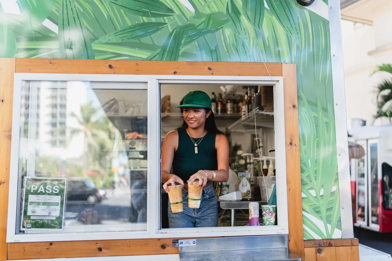 Usměvavá baristka asijského původu stojí za krásného slunečného odpoledne ve food trucku a podává z okénka dvě ledové kávy.