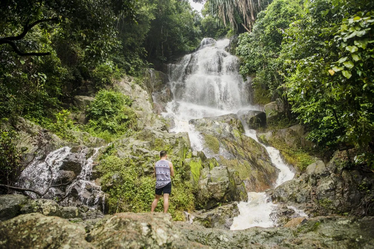 “Young man standing on rock looking at Na Muang Waterfall, Koh Samui, Thailand”