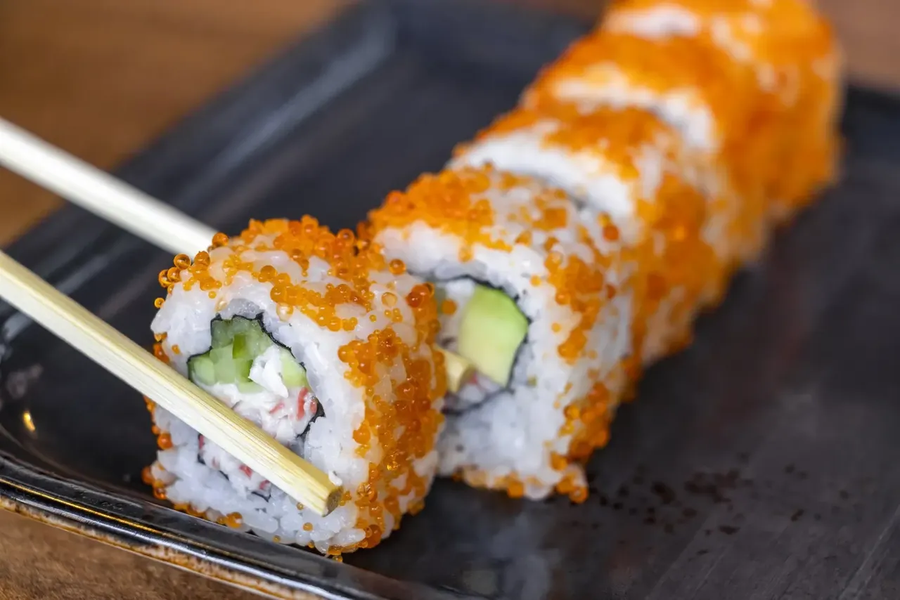 Un primer plano de rollos de sushi con tobiko de intenso color naranja, aguacate y surimi sobre un plato negro