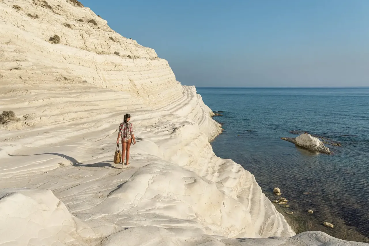 Woman with a straw bag wearing a shirt and swimwear walking on the white stone of the Scala dei Turchi. Realmonte, Agrigento, Sicily, Italy.