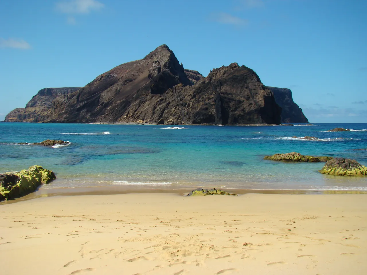Sandy beach at Calheta, Madeira with turquoise ocean water in the foreground and a rugged volcanic island rising from the sea under a clear blue sky.