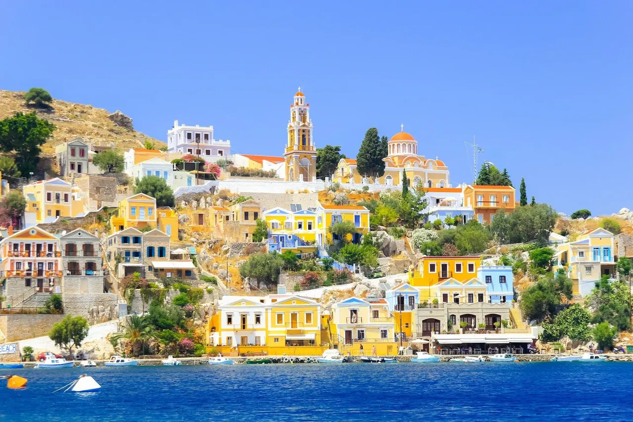 A stunning view of Simi Island, Greece, taken from a boat, showing colorful neoclassical buildings lining the harbor against a backdrop of rocky hills