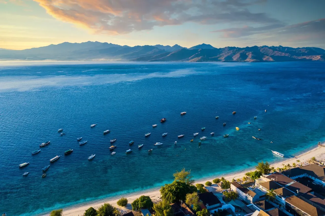 Toma aérea de la isla de Lombok vista desde Gili Trawangan, mostrando un tramo de costa con palmeras, playas y aguas cristalinas.
