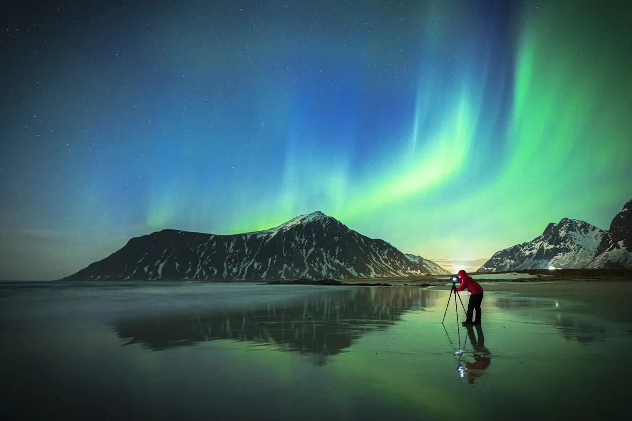 Fotograf ze statywem uwieczniający zorzę polarną na plaży Skagsanden, Flakstad na norweskich Lofotach