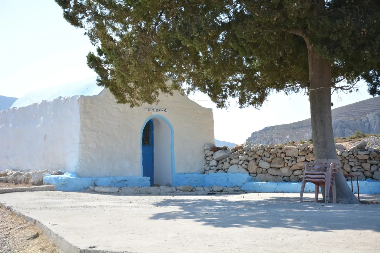 A quaint small white house in Tilos, Greece, featuring traditional Greek architecture with blue accents on doors and windows, surrounded by lush greenery
