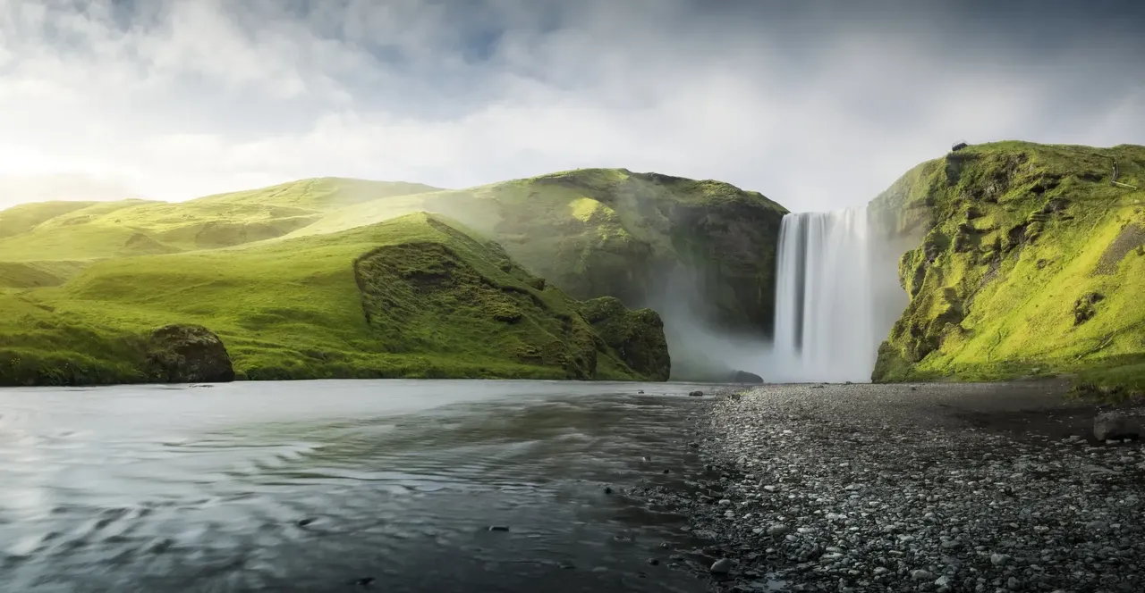 A Skógafoss vízesés egy zöldellő sziklafalon zúdul alá, előtte nyugodt folyó csordogál a völgyben