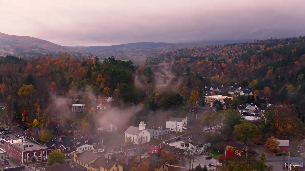 A misty aerial view of Stowe, Vermont, with rolling hills, colorful autumn foliage, and a serene fog blanketing the landscape.