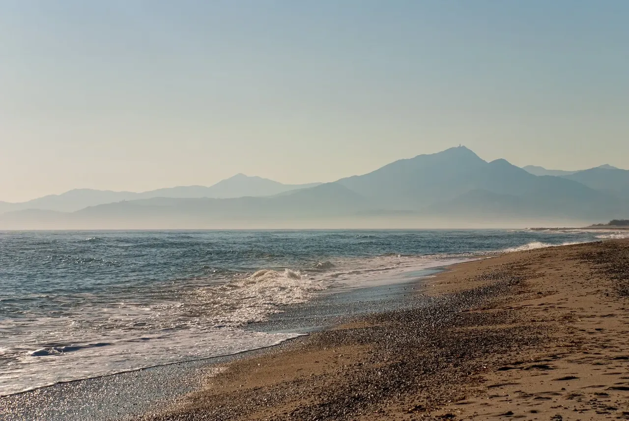 Foggy beach in wintertime in bright sunlight with the Albères mountain range in the background on a pale blue sky background, Argelès-sur-Mer, Céret, Occitanie, France, Europe.