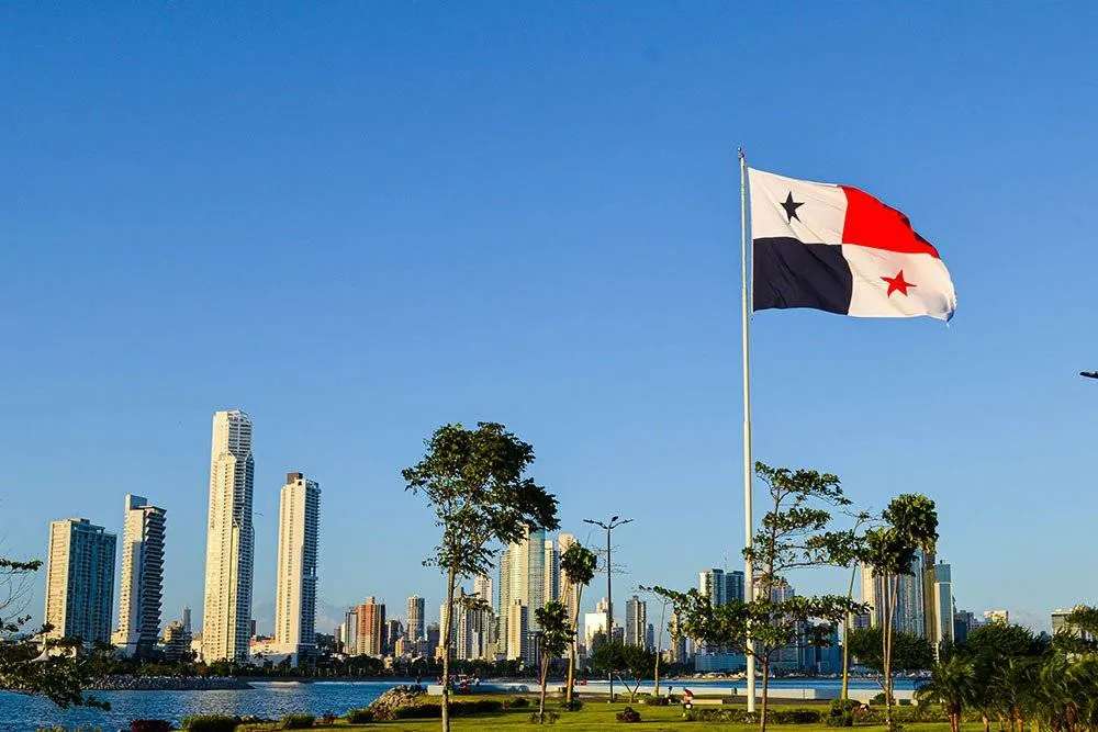 Panama zászlaja az árbócon, lobog a szélben, fűvel borított nyílt téren, a Csendes-óceán és a magas városi épületek mögül — Shutterstock