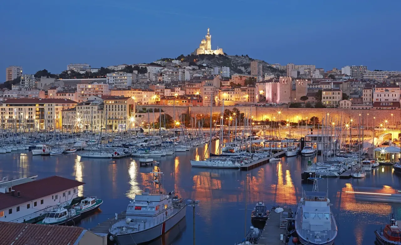 Kilátás a “Vieux Port” területére jachtokkal, a domb tetején lévő “Notre Dame de la Garde” éjszakai megvilágításban.