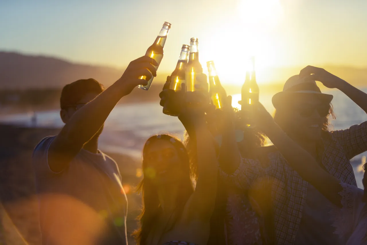Un grup de tineri petrecând pe plajă la apus. Toți au sticle de bere și sărbătoresc cu un toast. Siluete cu plaja, soarele și marea în fundal