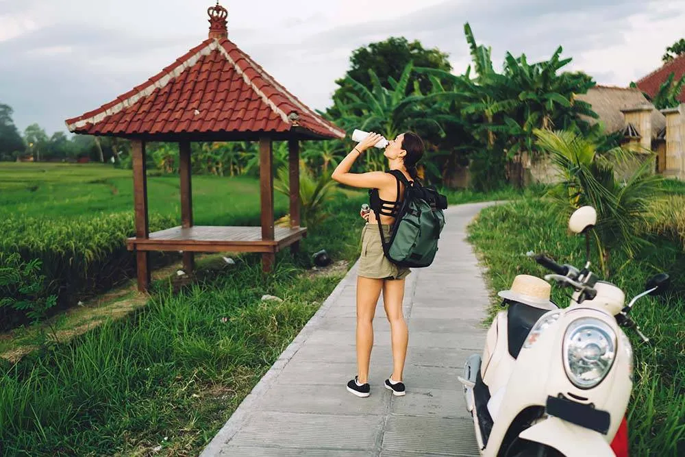 Back view of full body female traveler in casual outfit and backpack standing on path in rice field on Bali and drinking water