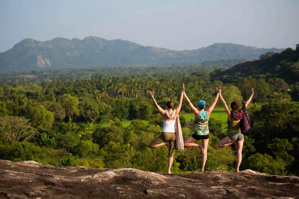 Three women making the same yoga pose on a rock with trees and hills in the background — ulpotha.com