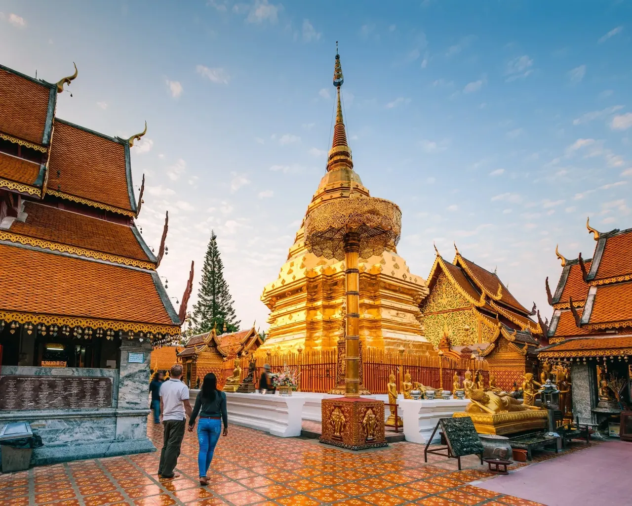 A majestic golden temple with intricate designs and a towering chedi, surrounded by lush greenery and overlooking the city of Chiang Mai from a mountain.