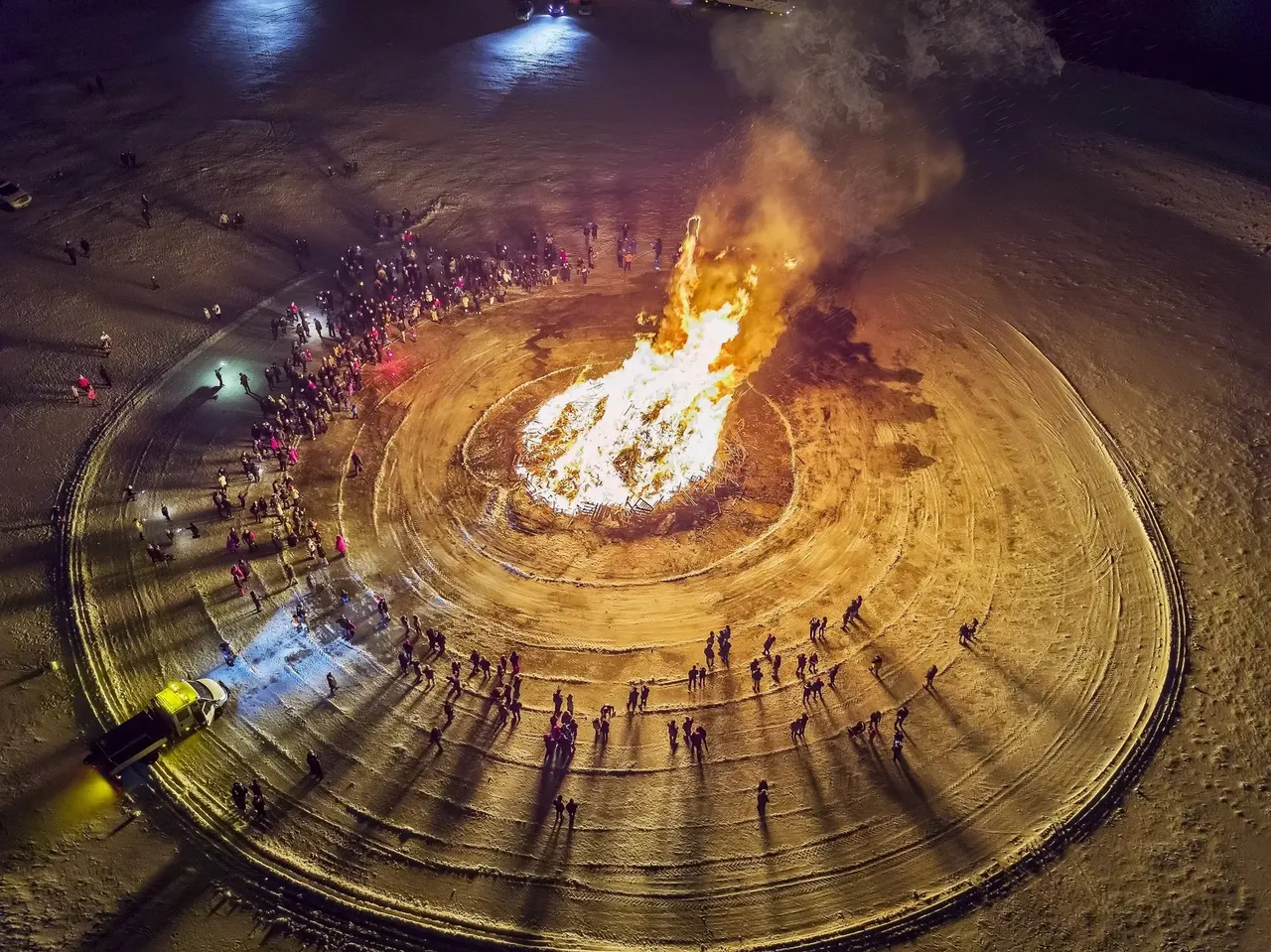 A massive bonfire surrounded by a crowd of people in a circular snow-covered area, viewed from above