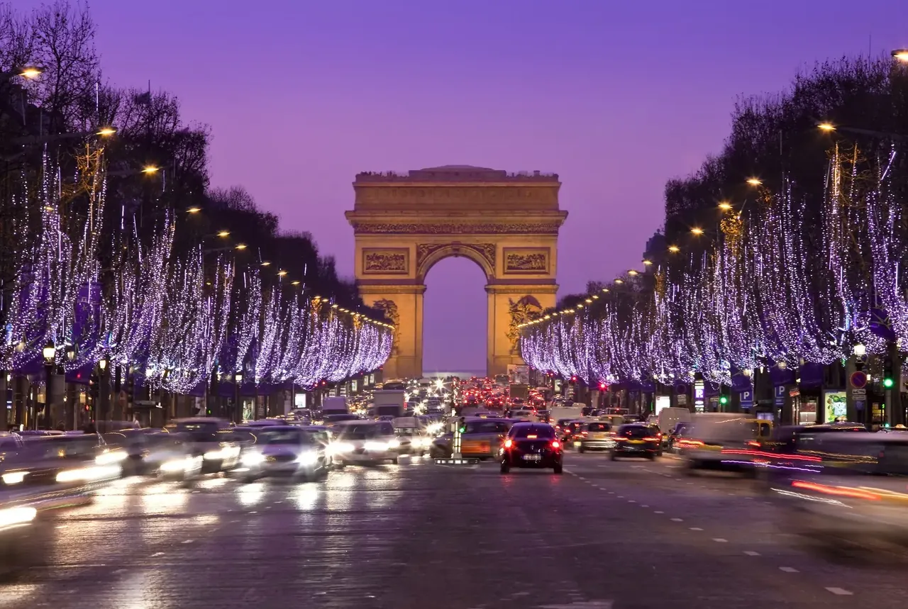 The Arc de Triomphe in Paris illuminated at night, framed by purple-lit trees lining the bustling Champs-Élysées