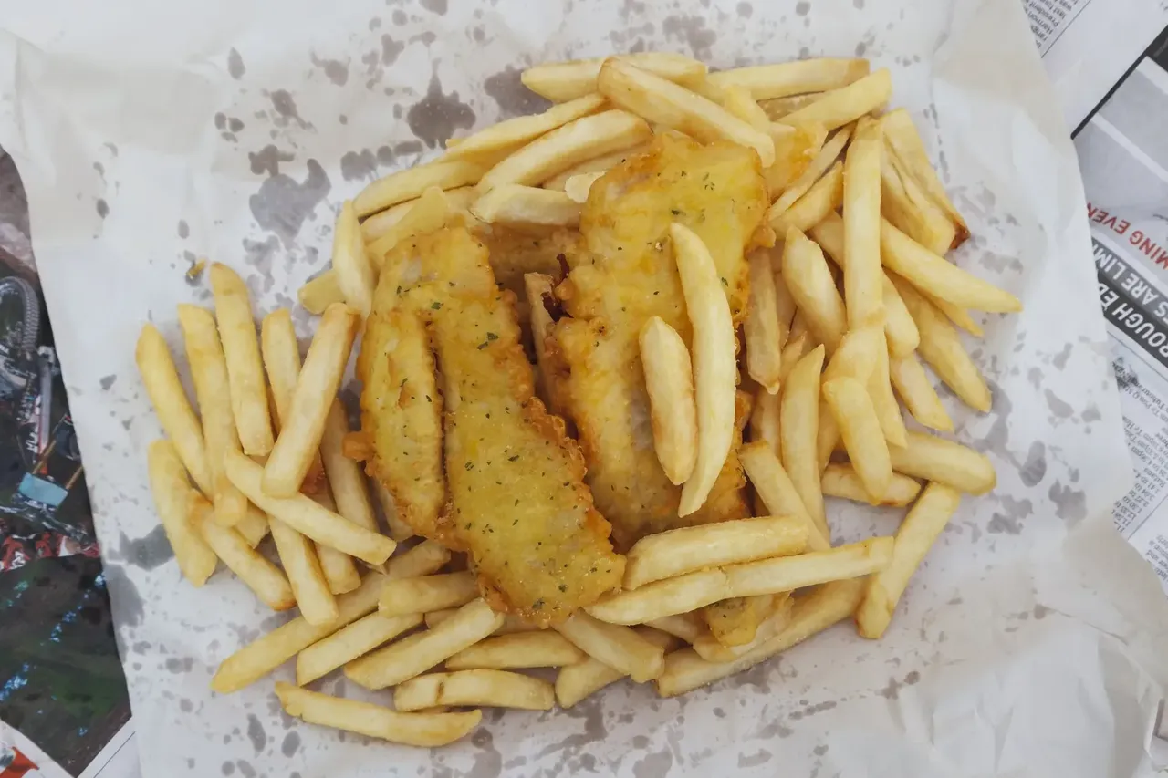 O vedere de aproape a unei mâncăruri britanice clasice de pește și cartofi prăjiți, cu fileuri de pește maroniu-auriu, înconjurate de cartofi prăjiți crocanți, servite pe o bucată de hârtie albă. Hârtia de dedesubt prezintă niște pete de grăsime, care sporesc senzația de autenticitate și dezinvoltură a acestei mese populare la pachet.