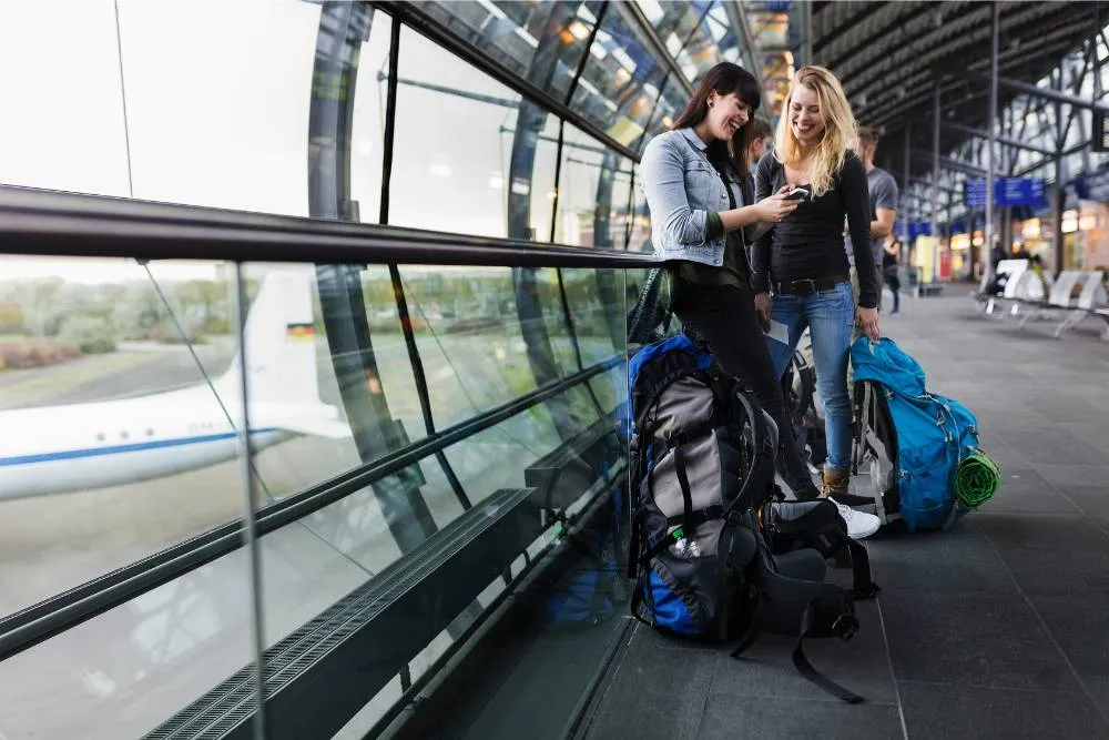Női hátizsákos utazók egy repülőtéren, telefonjukat nézegetik - Getty Images