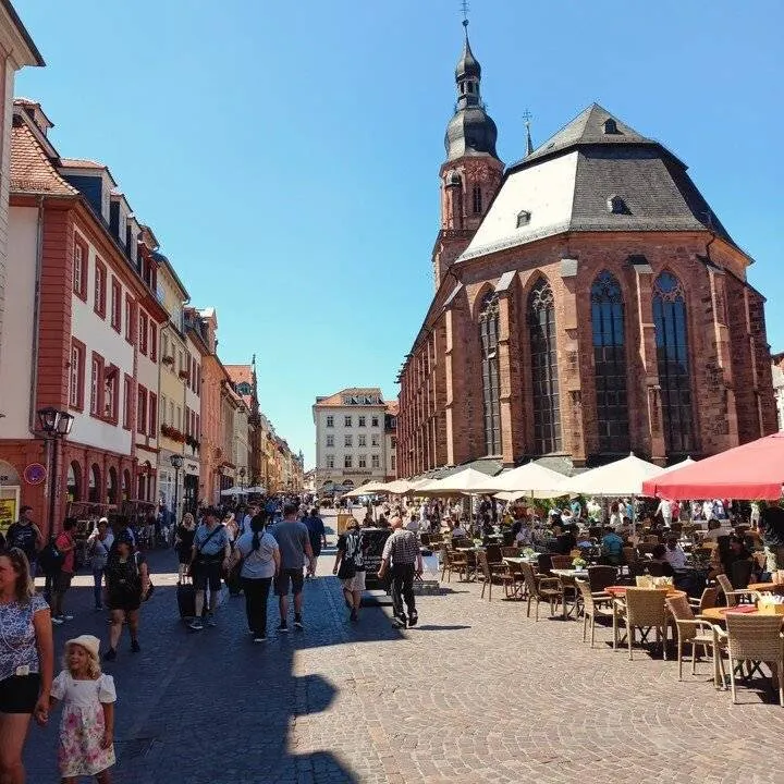 Heidelberg belvárosának nyüzsgő jelenete egy napsütéses napon, számos ember sétál a macskaköves utcán, körülötte vörös cseréptetős történelmi épületek, fölöttük tiszta kék ég.