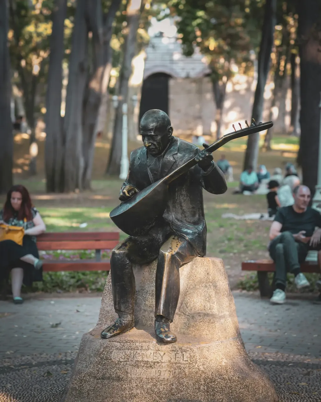 A bronze statue of a man playing a guitar, situated in a leafy park, surrounded by green trees and benches.