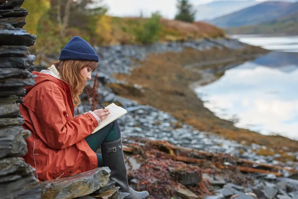 Woman sat by peaceful lake writing in journal — Getty Images