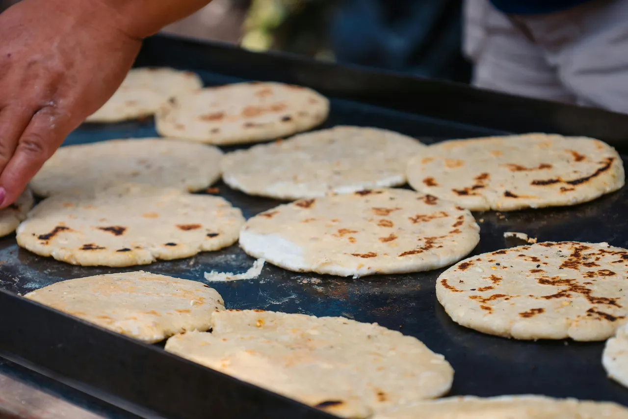 A man in Colombia grilling arepas on a metal grill, using a spatula to flip the golden corn cakes over an open flame.