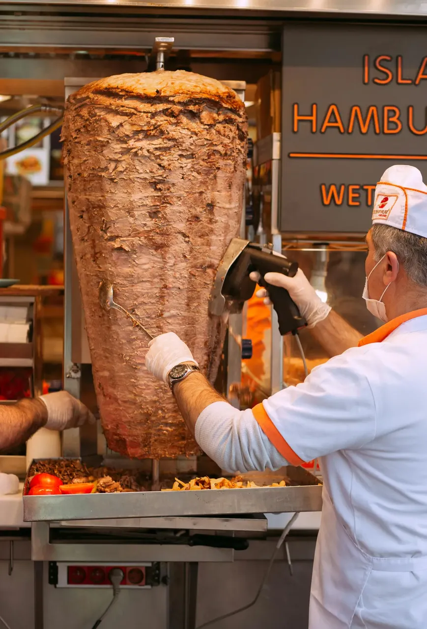 A man cutting slices from a large, vertical spit of doner kebab, with layers of marinated meat stacked in a conical shape, sizzling as it cooks.