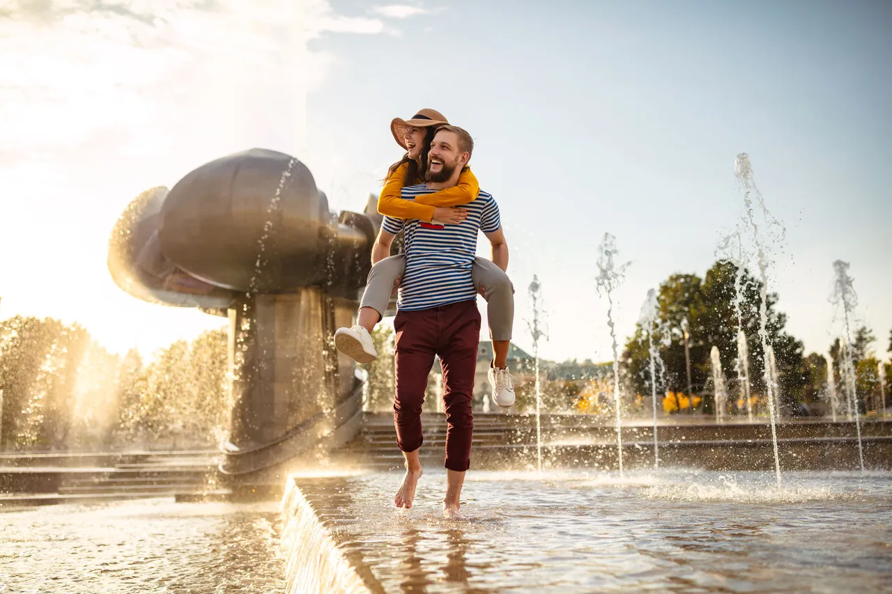 A couple enjoying a sunny day at the fountain on Námestie slobody in Bratislava with laughter and splashing water all around.