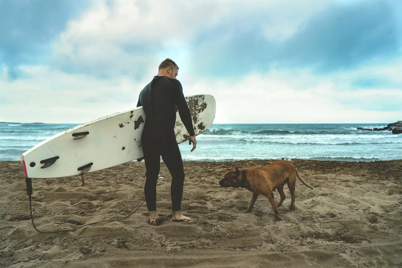 Man with a surf board standing on sandy beach with his big brown dog by the side