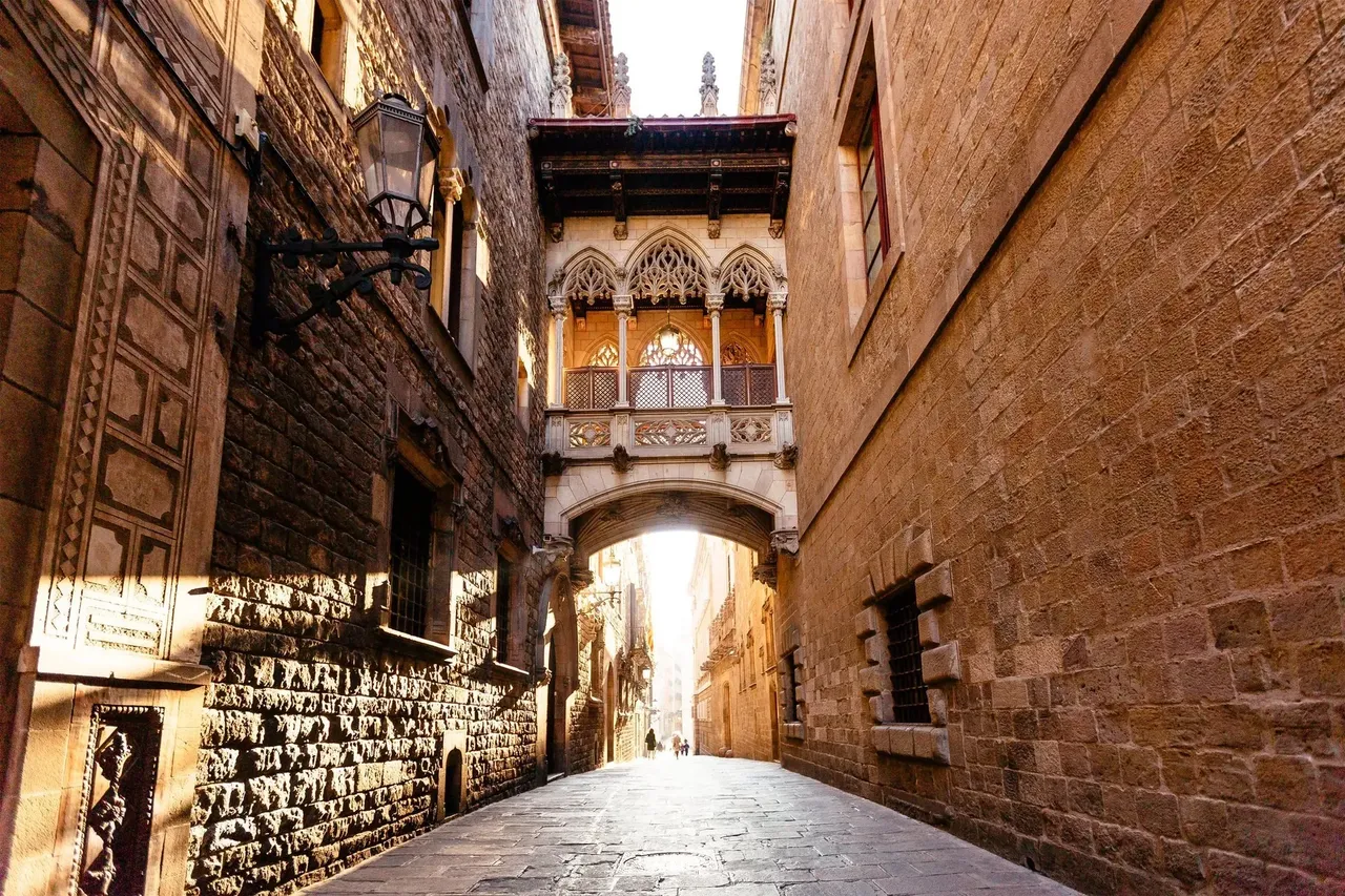 O stradă îngustă, cu piatră cubică, în Cartierul Gotic din Barcelona, mărginită de clădiri istorice, arcade și câțiva pietoni.