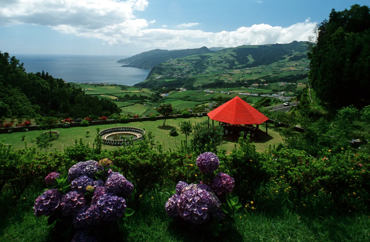 Scenic viewpoint in the Azores overlooking rolling green hills, patchwork farmland, and the Atlantic Ocean, with hydrangea flowers in the foreground and a red-roofed gazebo in a landscaped garden.