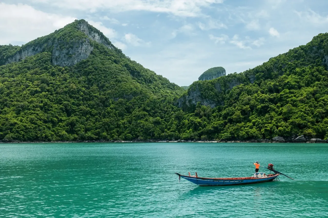 A picturesque scene of a man on a traditional wooden boat, floating on calm water with the backdrop of lush green mountains on Koh Samui.