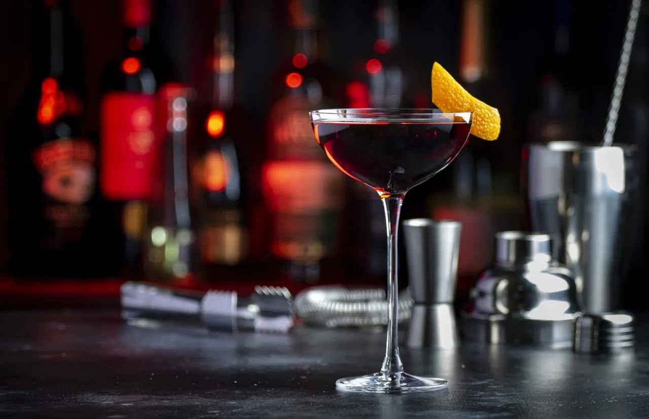 Adonis alcoholic cocktail drink with sherry and red vermouth, black bar counter background, steel tools and bottles