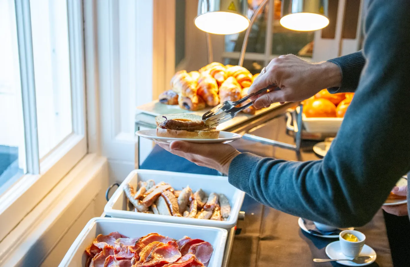 A guest serving themselves breakfast from a hotel buffet counter with bacon, sausages, and pastries