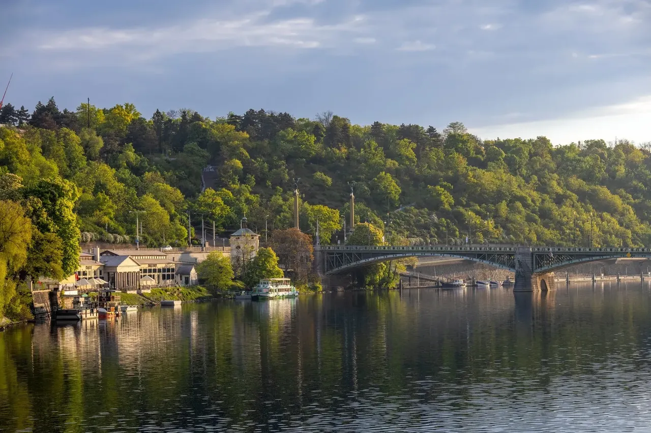 A calm view of the Vltava River, with a historic bridge and lush green hills in the background