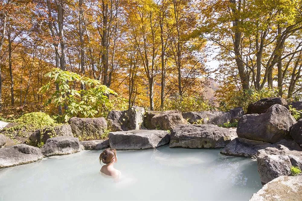Fürdőző nő egy szabadtéri japán fürdőben, a háttérben fák őszi lombkoronával
