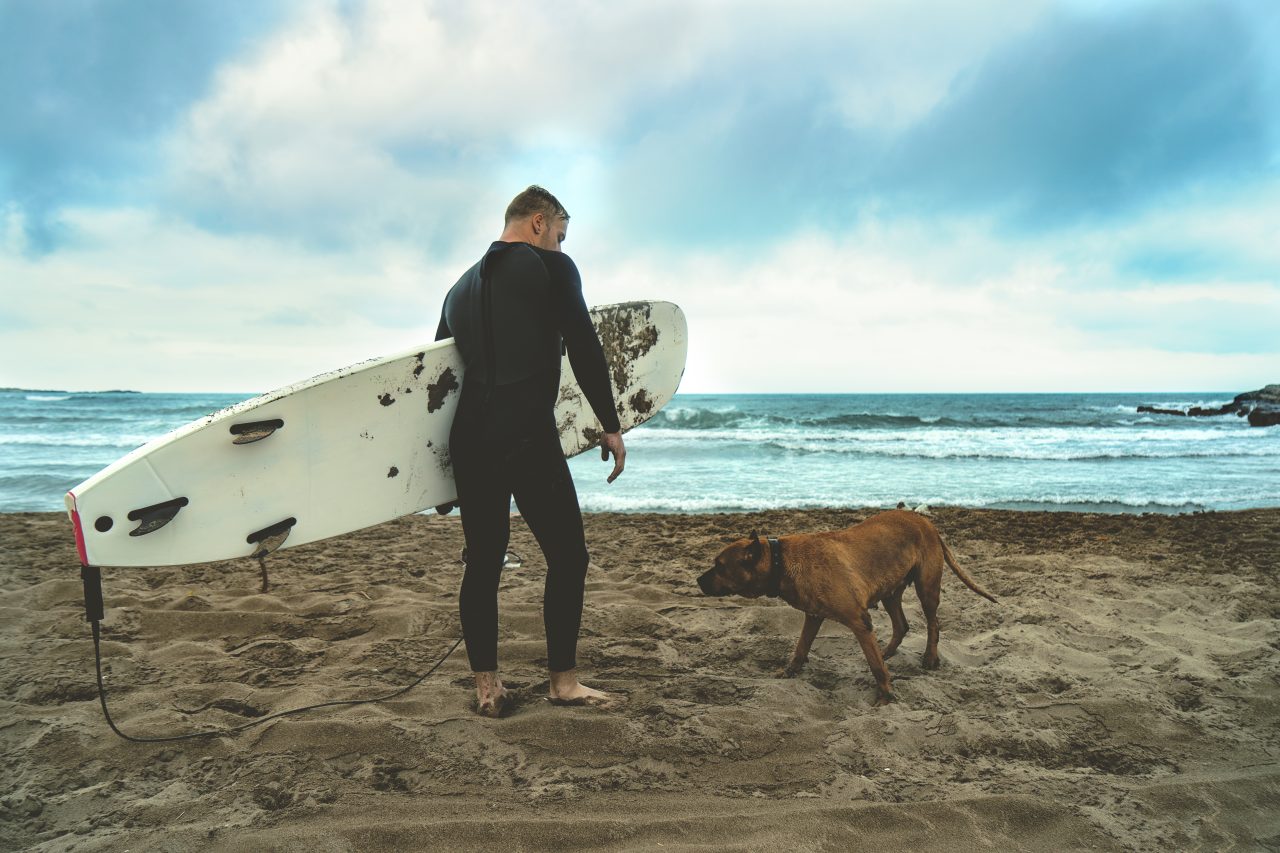 Man with a surf board standing on sandy beach with his big brown dog by the side 